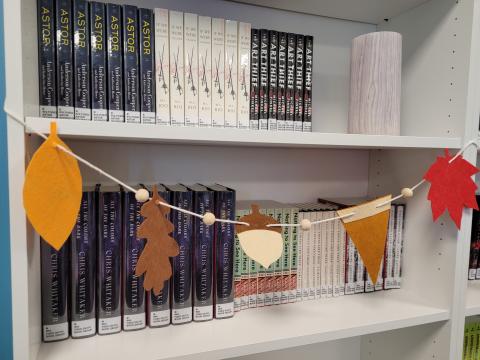 Image shows a white bookcase adorned with a string of garland featuring felt cutouts in the shape of fall colored leaves, an acorn, and a pumpkin pie slice, with small wooden beads between the cutouts. 