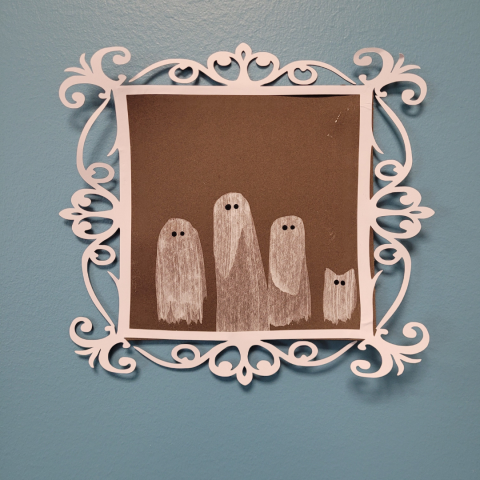 Image shows a square of dark brown paper with 3 ghost shapes made from a thin layer of white paint, with 2 black eye dots on each ghost. A small white ghost cat also appears to the right of the 3 ghost shapes. The ghost picture is adorned with a decorative cut-out white frame, and this is all against a blue background.
