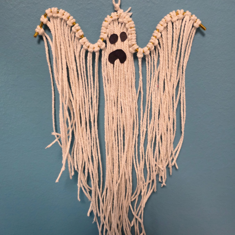 Image of a ghost made with macrame string hanging from a thin bent wire. Black construction paper is cut out and added to the string to make two eyes and a mouth.