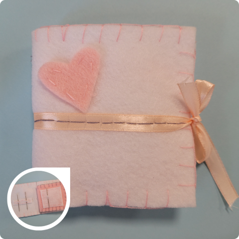 Image of a square felt booklet is displayed. The felt is white, with a pink felt heart hand-stitched to the front cover. Pink blanket stitching is along the booklet edges, and a light peach ribbon runs horizontally across the middle of the booklet cover and ties in a bow along the right side of the booklet to keep it closed. A close-up pop-out image shows the inside of the booklet, with two pages displaying sewing needles stored by being stuck through felt pieces on the pages.