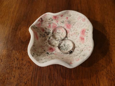 Image shows a white clay dish shaped like a small bowl with uneven wavy edges. The white dish has a sparse pink and green floral pattern inside, and three small rings are resting in the dish.