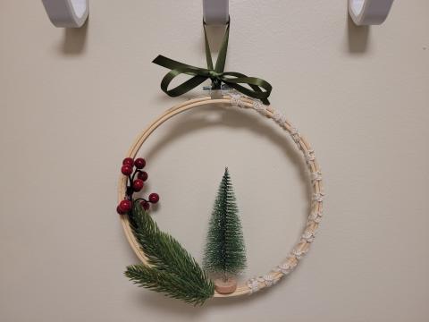 Image shows an embroidery hoop decorated with white lace ribbon wrapped around one half, a green bottle brush tree glued to the inside middle, a faux evergreen branch attached, and a sprig of red berries. The hoop is hung up on a white hook with a green ribbon bow tied at the top.