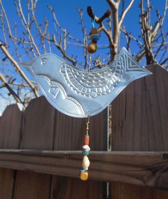 Silver aluminum in shape of bird with beads hanging down in front of brown fence, tree branches, and blue sky.